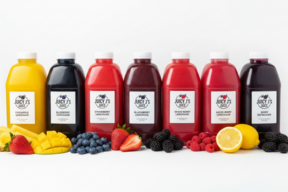 Eight bottles of Juicy's fruit smoothies arranged in a row on a white background, with corresponding fresh fruits placed in front of each bottle. The bottles range from yellow and orange to red and dark purple/black, and the labels clearly show the Juicy's brand.